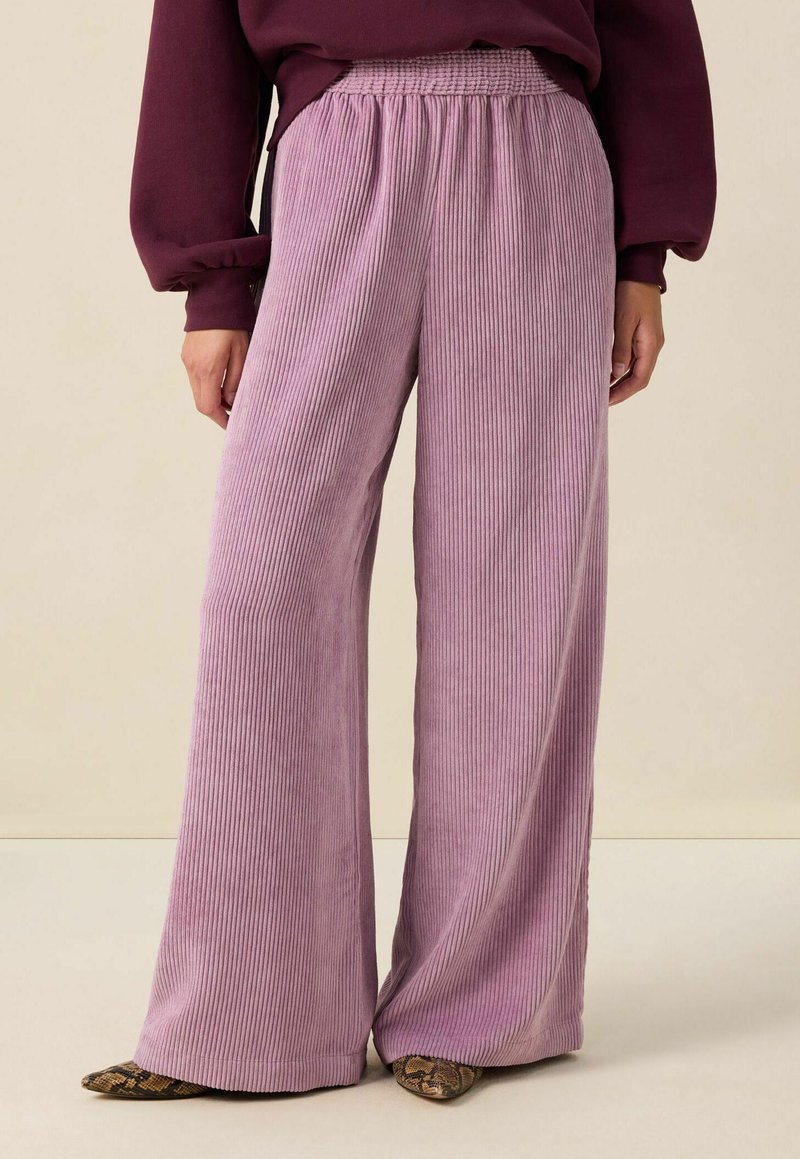 Pantalons larges en tissu lilas côtelé et doux. Présentent une taille élastique et une silhouette fluide, associés à des chaussures à motifs sombres.