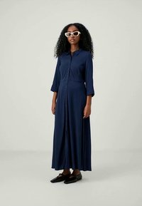 Jūras zilas krāsas, pogu aizdare maxi kleita ar trim ceturtdaļa piedurknēm un piegulošu vidukli, ar gludu, plūstošu audumu un minimālistisku dizainu.