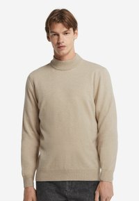 Junger Mann, der einen beigen Rollkragenpullover und dunkelgraue Jeans vor einem schlichten weißen Hintergrund trägt.