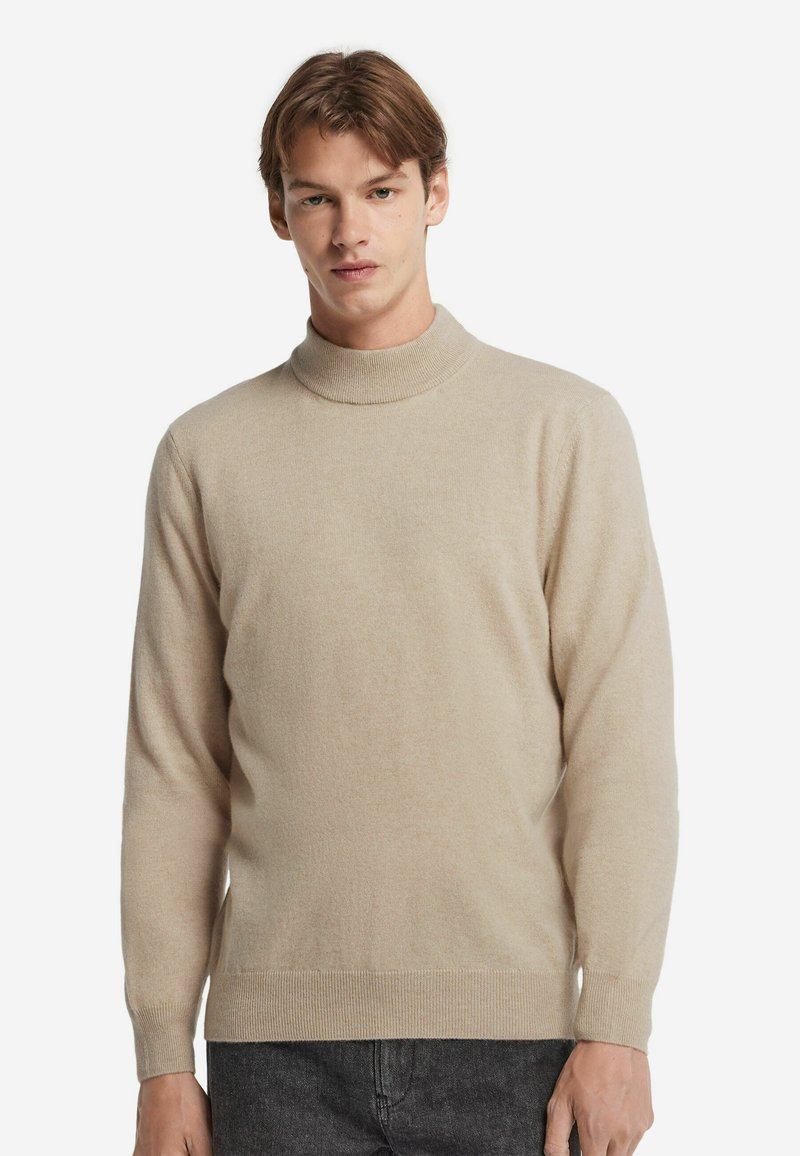 Junger Mann, der einen beigen Rollkragenpullover und dunkelgraue Jeans vor einem schlichten weißen Hintergrund trägt.