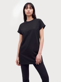 Mujer con el pelo liso y oscuro, que lleva un vestido suelto tipo camiseta negra de manga corta y pantalones negros, de pie frente a un fondo claro y sencillo.