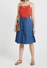 Camiseta roja sin mangas con encaje, falda de mezclilla en forma de A con botones frontales, sandalias de cuero marrón y bolsa cruzada de mimbre.