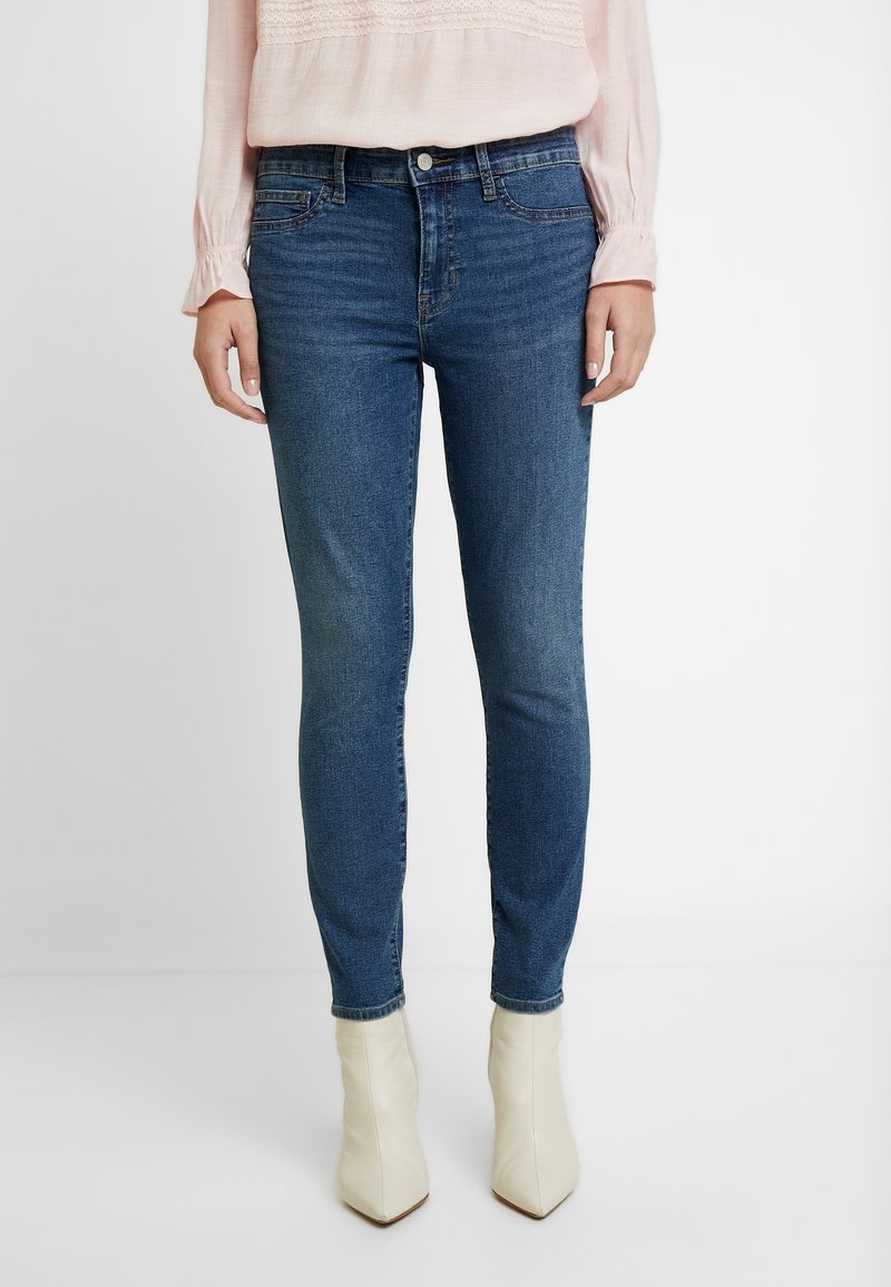 Femme portant un jean skinny bleu foncé, un chemisier rose clair à manches longues et des bottines blanches à bouts pointus, debout sur un fond blanc.