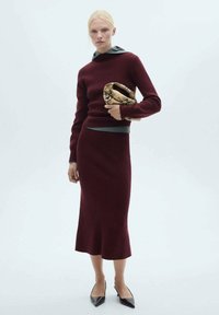 Maglione lavorato a maglia bordeaux con cappuccio grigio, abbinato a una gonna midi coordinata. Tessuto testurizzato, silhouette aderente e tacchi a punta neri.