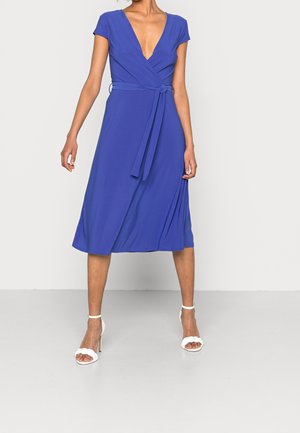 Femme portant une robe portefeuille bleu roi mi-longue à manches courtes et des sandales blanches à talons hauts, debout sur un fond clair uni.