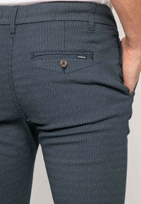 Pantalon bleu marine avec un motif en diamant texturé, une fermeture de poche arrière standard et une petite étiquette de marque au-dessus de la poche.