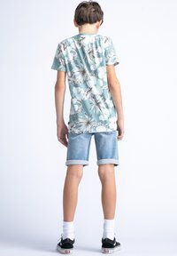 Camisa de manga corta en teal claro con grandes patrones de flores blancas y verdes, combinada con pantalones cortos de mezclilla arremangados y zapatillas negras.