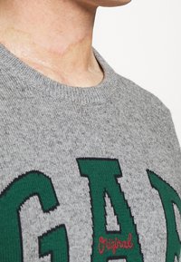 Close-up van de nek van een persoon en een grijze gebreide trui met grote groene letters "GAP" en kleine rode geborduurde tekst "Original".