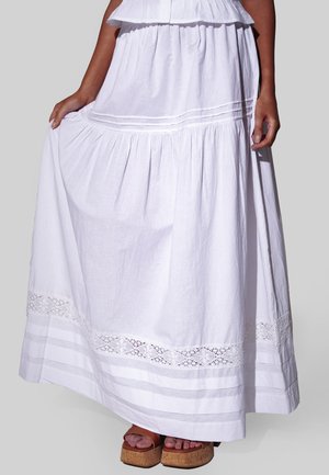 Vrouw draagt een lange witte laagjesrok met kanten rand, houdt de stof vast, gecombineerd met bruine plateauzandalen.