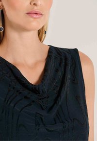 Donker marineblauw mouwloos topje met een gedrapeerde halslijn, voorzien van gestructureerde golfpatronen en een lichte glans. Gecombineerd met lange oorhangers.