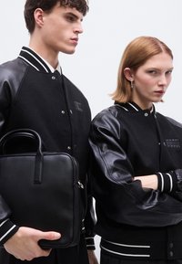 Jachete bomber negre cu corpuri din lână și mâneci din piele, având manșete striate și un buzunar cu fermoar, asortate cu o geantă neagră din piele.