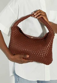 Brown woven handbag with a curved handle, textured surface, and subtle sheen. Features a magnetic closure and sits against a white shirt.