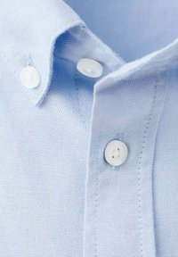 Gros plan sur une chemise bleu clair, présentant un tissu texturé, un col pointu et des boutons en plastique blanc. Coutures visibles le long des bords.