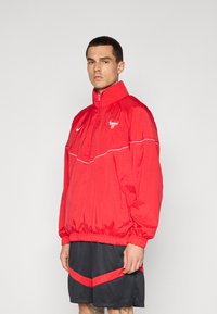 Rød vindtæt jakke med høj krave, elastiske manchetter og talje. Har et logo på brystet og hvide accentlinjer. Sammen med sorte shorts.