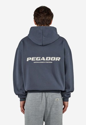 Osoba nosząca ciemną bluzę z kapturem z napisem "PEGADOR Certified Authentic Streetwear" na plecach, w połączeniu z jasnoszarymi, fakturowanymi spodniami.