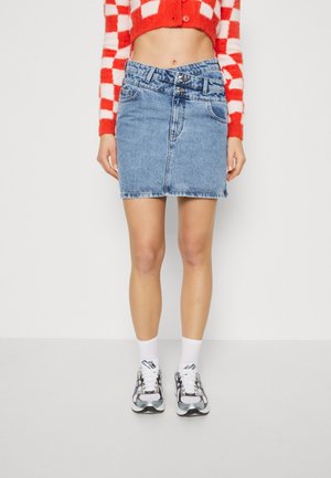 Kvinde iført en blå denim mininederdel med dobbelt knap i taljen, rød- og hvidternet kort sweater, hvide sokker og grå sneakers.