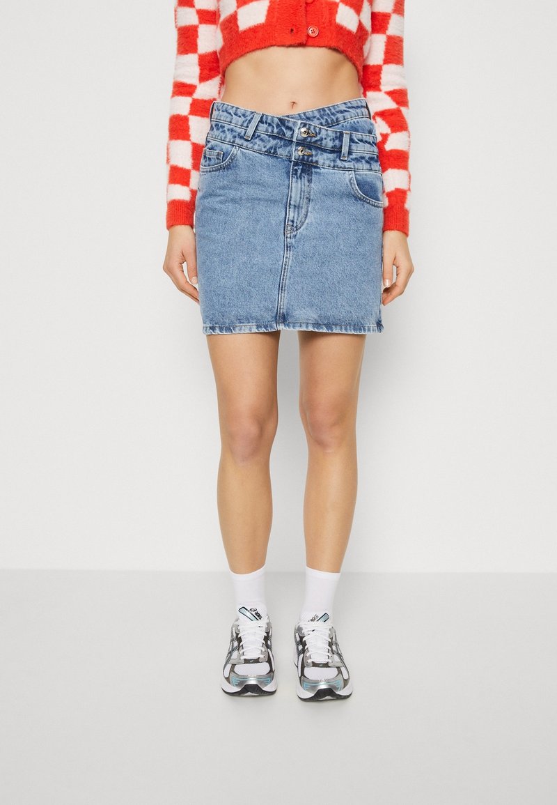 Femme portant une mini-jupe en jean bleu avec une double boutonnière à la taille, un pull court à carreaux rouges et blancs, des chaussettes blanches et des baskets grises.