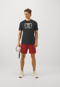 Schwarzes Kurzarmhemd mit einem silbernen Under Armour-Logo, kombiniert mit roten Shorts; hält ein Springseil; beiges Sneakers, glatte Textur.