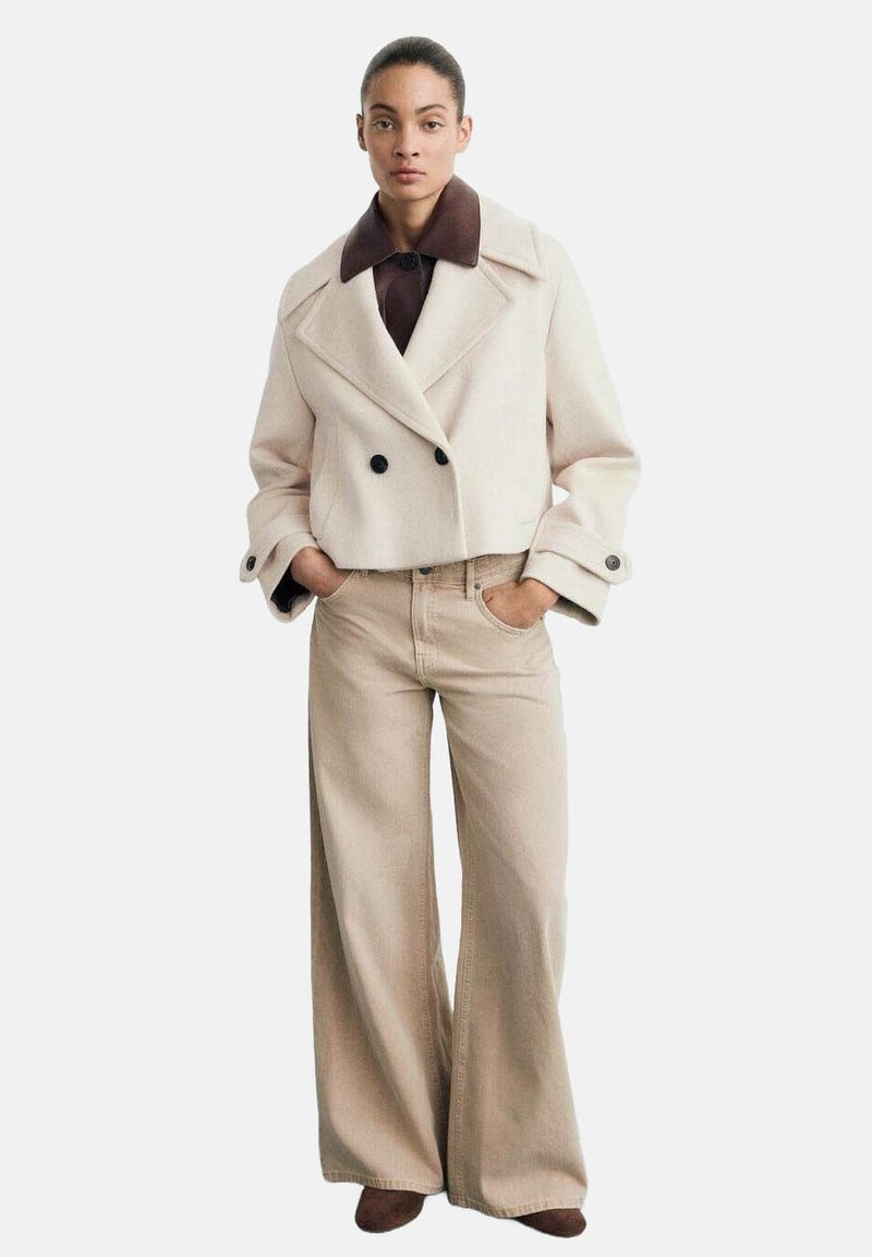 Kremowy, podwójnie zapinany płaszcz wełniany z brązowym kołnierzem i ciemnymi guzikami, w zestawieniu z jasnobeżowymi spodniami szerokimi w nogawkach.