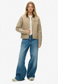 Veste en coton beige avec un col foncé, fermeture éclair et poche avant, associée à un jean large bleu clair et des baskets beiges.