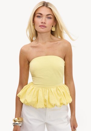 Femme aux longs cheveux blonds portant un top jaune bustier à basque, un pantalon blanc, des boucles d'oreilles dorées et de gros bracelets en or et argent.