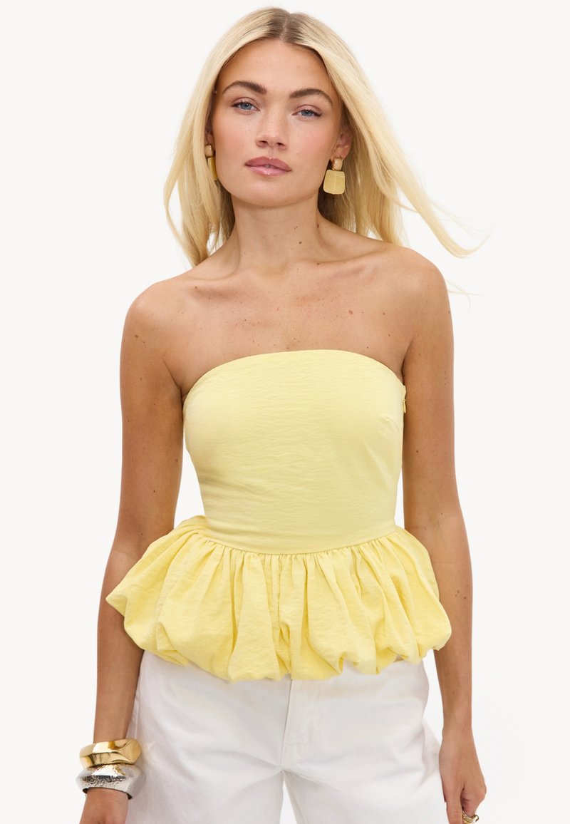 Femme aux longs cheveux blonds portant un top jaune bustier à basque, un pantalon blanc, des boucles d'oreilles dorées et de gros bracelets en or et argent.