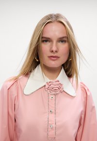 Blusa rosa con colletto bianco, caratterizzata da un'applicazione a forma di rosa, decorazioni in strass e maniche a sbuffo. Tessuto morbido e vestibilità sartoriale.