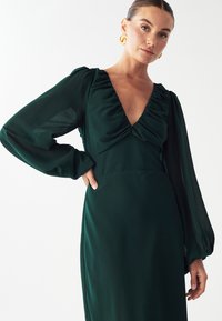 Zelené šaty s výstrihom do V, volánkovým telíčkom a dlhými priehľadnými rukávmi. Hladká látka sa jemne splýva, pričom zvýrazňuje štíhlu, elegantnú siluetu.
