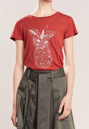 Persoon draagt een roestkleurig T-shirt met een witte ananasschets en een hoge taille olijfgroene rok met riem.