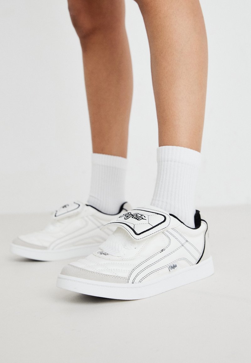 Sapatilhas brancas de desporto com um exterior sintético liso, detalhes pretos e fecho de velcro. Apresentam meias brancas com relevo e um design de sola simples.