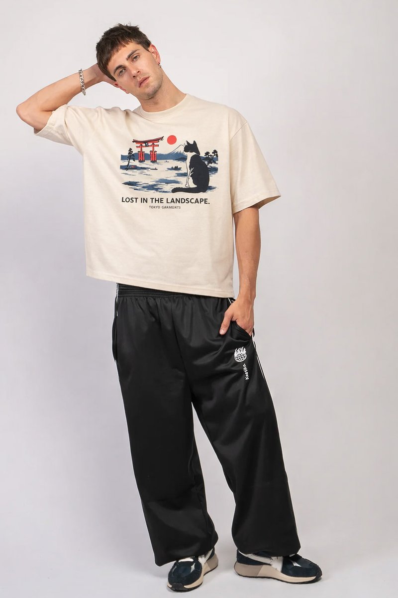 Young man posing with hand behind head wearing beige graphic T-shirt and loose black pants against plain background.