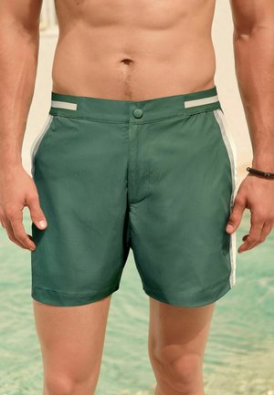 Grüne Herren-Badeshorts mit weißen Seitenstreifen, vorne mit Knöpfen, getragen von einem oberkörperfreien Mann, der in der Nähe von Wasser steht.
