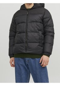 Sort dunjakke med hætte, som har lynlås, quiltet design og sidelomme. Båret over en grøn striktrøje og denimjeans.