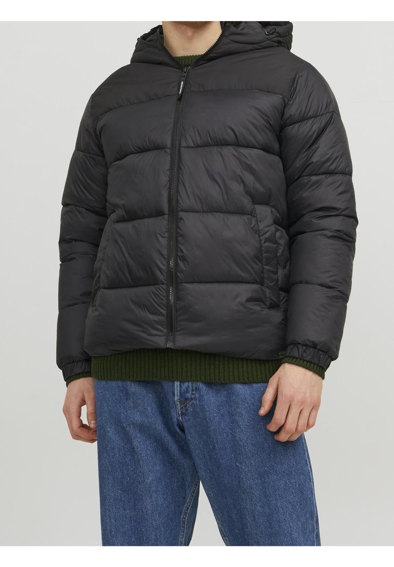 Sort dunjakke med hætte, som har lynlås, quiltet design og sidelomme. Båret over en grøn striktrøje og denimjeans.