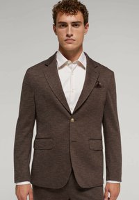 Hnedý blazer v herringbone vzore s klopovými goliermi, dvoma prednými vreckami a jedným gombíkom na zapínanie. Na tričku bielej farby.