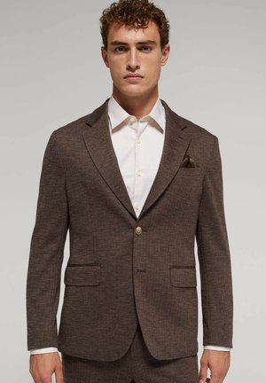 Blazer marrone a losanga con rever a profilo, due tasche frontali e chiusura con un solo pulsante. Indossato sopra una camicia bianca.