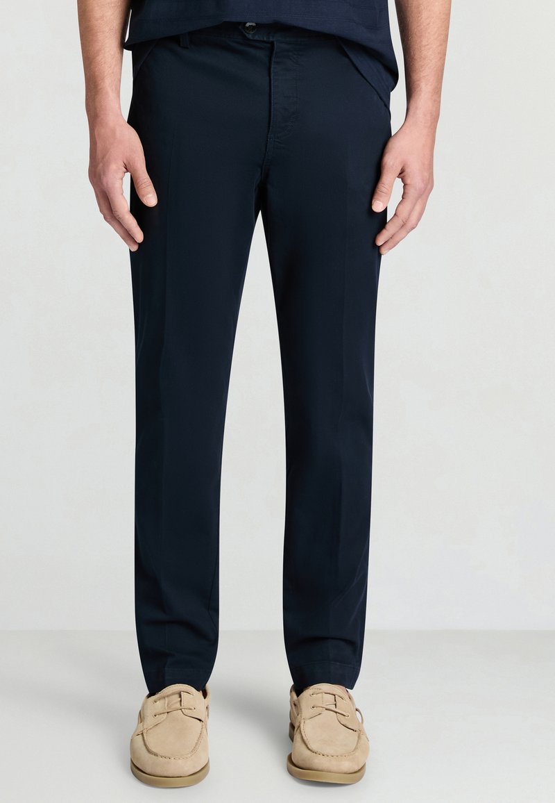 Marineblaue Chinos mit flacher Vorderseite, aus einem glatten Stoff gefertigt, mit Seitentaschen und klassischem Schnitt. Kombiniert mit beigen Slippern.