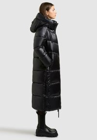 Lange zwarte pufferjas, glanzende afwerking met een capuchon, ritsluiting, zijzakken en een boxy silhouet. Gekozen in combinatie met zwarte chunky laarzen.