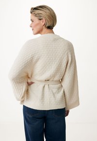 Krämfärgad, texturerad kappa med öppen front, vida ärmar och matchande bälte, parat med mörkblå jeans mot en ljus bakgrund.