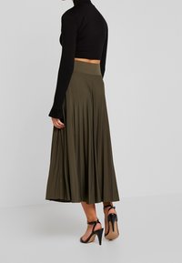 Olive green pleated maxi skirt with a smooth texture and high waist, paired with a black cropped long-sleeve top and black high-heeled sandals.