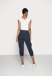 Wit top met kant, marineblauwe cropped broek met elastische taille, en witte sneakers met zwarte accenten, van achteren bekeken tegen een effen achtergrond.