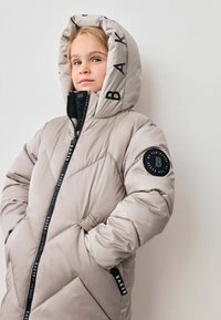 Giacca imbottita per bambini color beige chiaro con motivo trapuntato, cappuccio alto, dettagli in logo neri, chiusura con zip e tasche laterali.