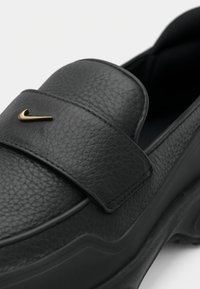 Gros plan d’une chaussure noire en cuir texturé à enfiler avec une semelle épaisse et un petit logo doré Nike sur la bride supérieure.