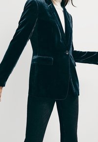 Femme portant un blazer en velours sombre ajusté avec un seul bouton et un pantalon assorti de couleur foncée, sur un fond clair uni.