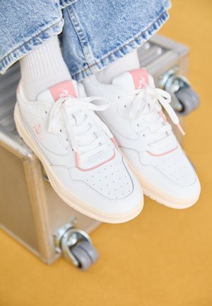 White leather sneakers with pink accents, perforated toe, flat laces, and a soft, textured sole, placed on a metallic surface.