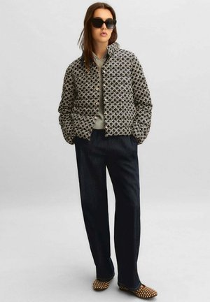 Donna che indossa una giacca trapuntata nera con motivo, felpa con cappuccio grigia, pantaloni ampi scuri, ballerine marroni borchiate e grandi occhiali da sole neri, in piedi con le mani nelle tasche.