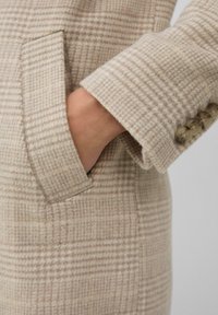 Hand in de zak van een beige en crème geruite wollen jas met vier knopen op de manchet tegen een neutrale achtergrond.