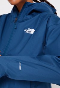 Personne portant une veste bleue North Face avec une fermeture éclair frontale et une étiquette en tissu dryvent sur la manche.