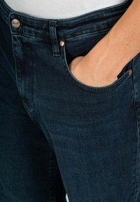 Los jeans de mezclilla azul oscuro presentan un diseño clásico de cinco bolsillos con una mano visible en el bolsillo frontal. Acentos de botones metálicos en la cintura.