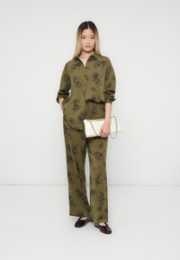 Donna che indossa un completo di pantaloni con camicia a stampa floreale verde oliva, mentre tiene una borsa bianca con tracolla a catena, in piedi su uno sfondo bianco uniforme.